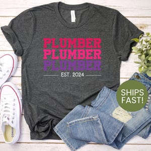 May include: A dark gray t-shirt with the text "PLUMBER" repeated three times in pink, purple, and red. The text "EST. 2024" is below the repeated text.