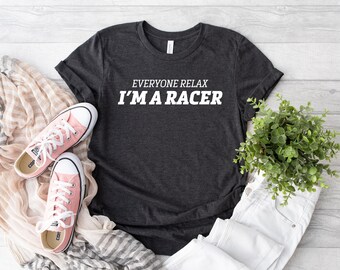 Puede incluir: Una camiseta gris oscuro de manga corta con el texto blanco "EVERYONE RELAX I'M A RACER" impreso en el pecho. La camiseta se muestra junto a un par de zapatillas de lona rosa claro, una bufanda beige estampada y una pequeña planta verde.