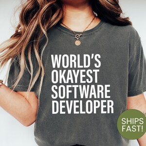 May include: A charcoal grey short-sleeve t-shirt featuring the white text "WORLD'S OKAYEST SOFTWARE DEVELOPER" printed in a bold, sans-serif font across the chest. The garment has a crew neckline and a relaxed fit, made of a solid-coloured fabric.