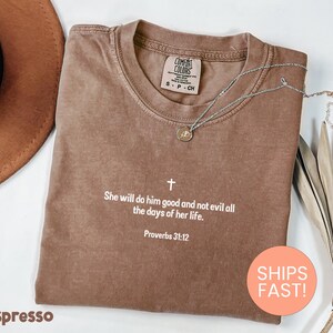 May include: A brown short-sleeved t-shirt with a crew neckline featuring a white cross icon and the text "She will do him good and not evil all the days of her life. Proverbs 31:12" printed in white on the centre chest area of the garment.