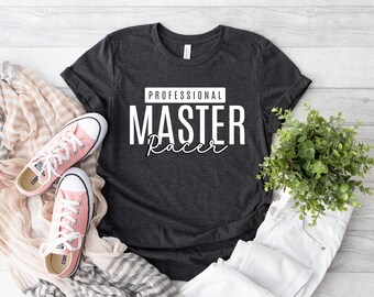 Puede incluir: Una camiseta gris oscuro de manga corta con el texto blanco PROFESSIONAL MASTER Racer impreso en el frente. La prenda se muestra junto a un par de zapatillas de lona rosa claro, una bufanda a rayas beige y blanca y una pequeña planta verde.