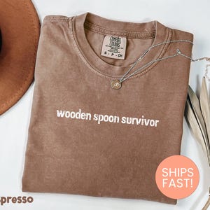 May include: Espresso-colored t-shirt with the text "wooden spoon survivor" in white. A silver necklace with a pendant rests on the shirt. The shirt is folded and displayed with a hat and dried plant.