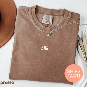 May include: Espresso-colored t-shirt with the word "bibi" in white lettering. A silver necklace with a pendant rests on the shirt. The shirt has a Comfort Colors tag. A round sticker with "SHIPS FAST!" is visible.