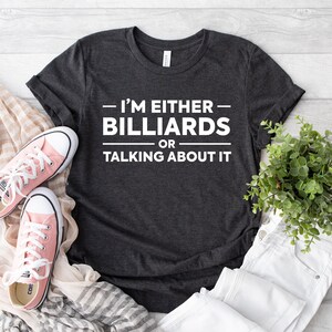 May include: A dark charcoal gray short-sleeved t-shirt featuring the white text "I'M EITHER BILLIARDS OR TALKING ABOUT IT" centered on the chest. The shirt is displayed alongside a pair of light pink canvas sneakers and a small green potted plant.
