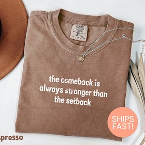 May include: Espresso-colored t-shirt with the text "the comeback is always stronger than the setback" in white. A silver necklace with a pendant rests on the shirt. The shirt is folded, with a brown hat and dried leaves in the background.