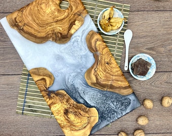 Puede incluir: Tabla de servir de madera hecha a mano con un diseño de río de resina azul claro. La tabla presenta vetas de madera natural y una forma rectangular. Una taza de café, higos secos y nueces están dispuestas sobre una mesa de madera.