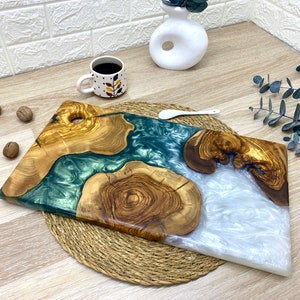 May include: Two wooden serving boards with epoxy resin accents. One board features a teal resin river, the other a white resin river. The boards are made of olive wood and are displayed on a woven mat.