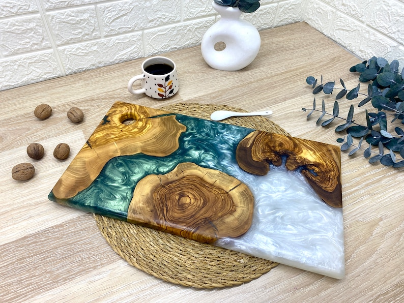 May include: Two wooden serving boards with epoxy resin accents. One board features a teal resin river, the other a white resin river. The boards are made of olive wood and are displayed on a woven mat.