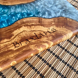 May include: A wooden serving board with a river of blue and silver resin. The wood has a natural grain pattern and the name "Braxtaris" is etched into the wood. The board is on a bamboo mat.