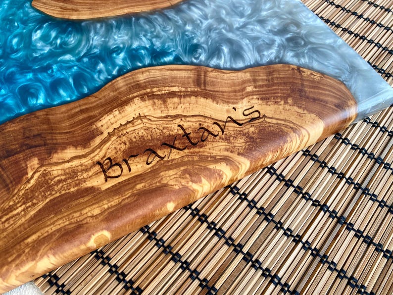 May include: A wooden serving board with a river of blue and silver resin. The wood has a natural grain pattern and the name "Braxtaris" is etched into the wood. The board is on a bamboo mat.