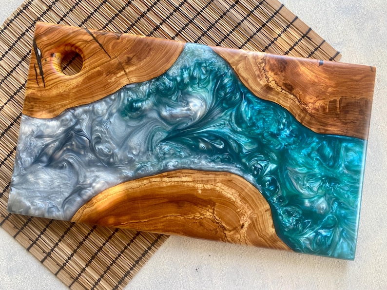 May include: A rectangular wooden serving board with a river of teal and silver resin. The wood has a natural grain and a hole for hanging. The resin flows through the center, creating a unique and decorative design. This board is perfect for serving appetizers or as a decorative piece.