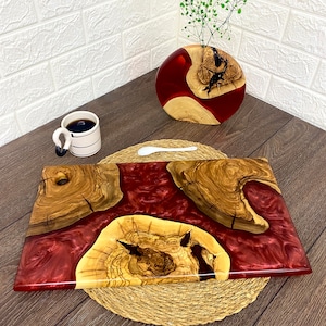 May include: A handcrafted serving board with a deep red resin center and olive wood accents. The board is rectangular, with a natural wood grain pattern. A matching vase with a similar design and a cup of coffee are also present.