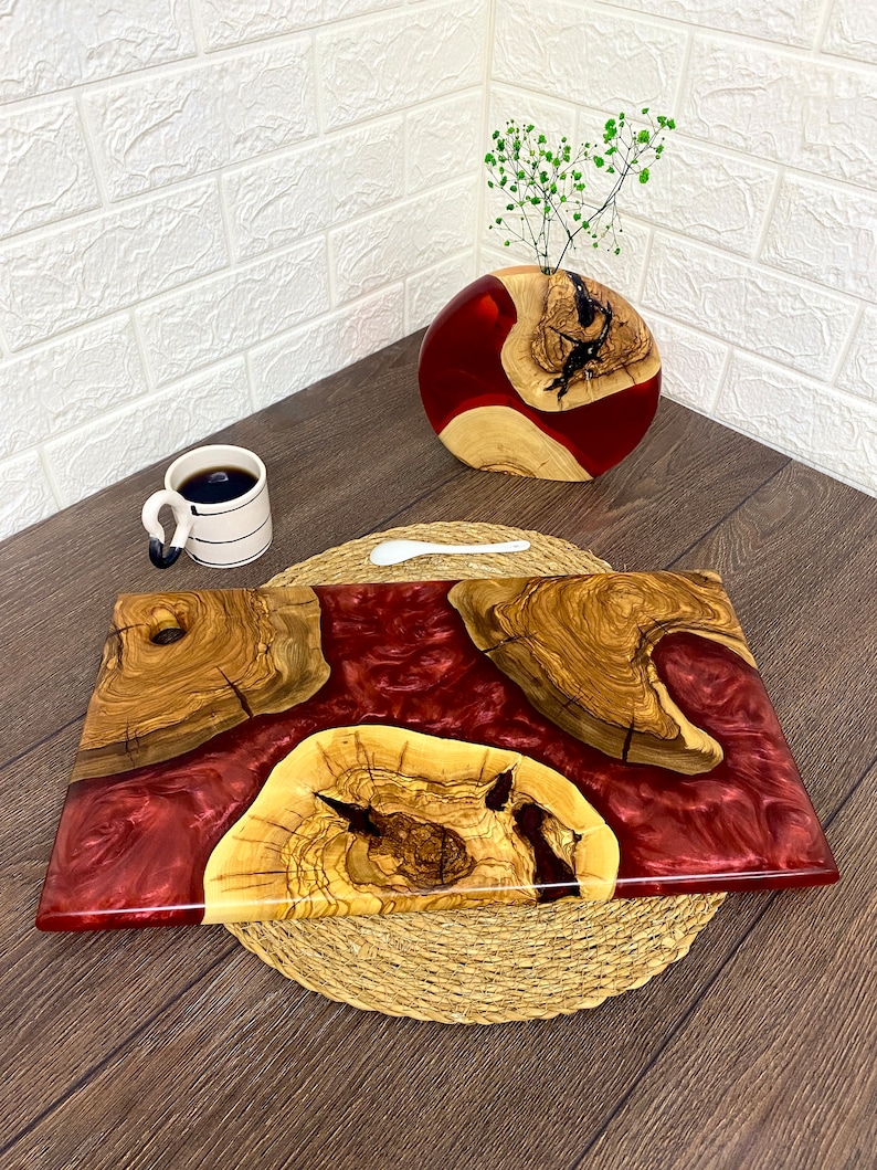 May include: A handcrafted serving board with a deep red resin center and olive wood accents. The board is rectangular, with a natural wood grain pattern. A matching vase with a similar design and a cup of coffee are also present.