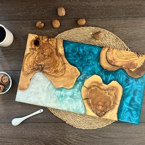 May include: A handcrafted wooden serving board with a turquoise resin river design. The board features natural wood grain and a heart-shaped wood element. A cup of coffee, small bowl, spoon, and walnuts are also present.