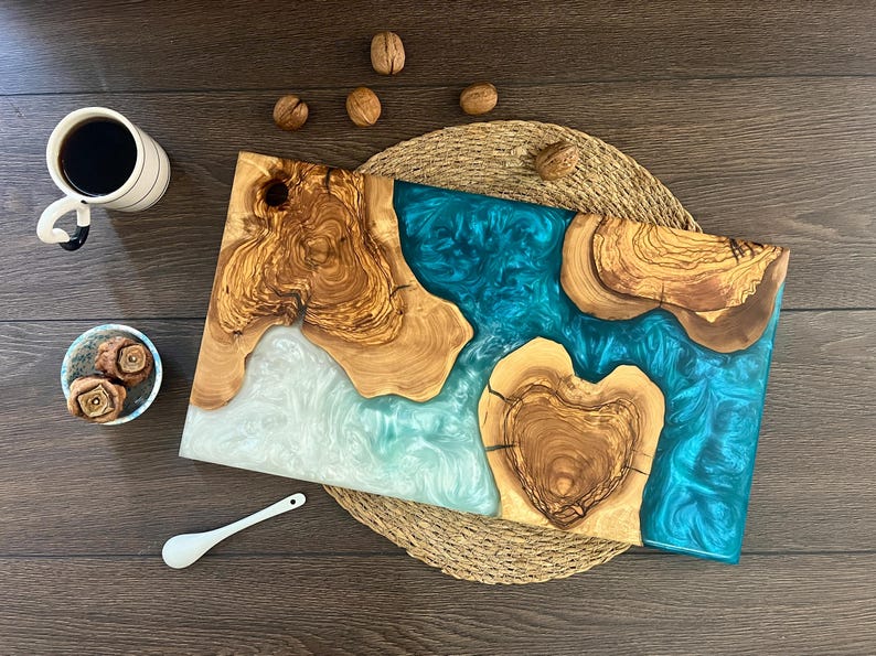 May include: A handcrafted wooden serving board with a turquoise resin river design. The board features natural wood grain and a heart-shaped wood element. A cup of coffee, small bowl, spoon, and walnuts are also present.