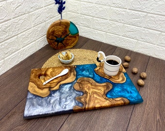 Puede incluir: Una bandeja de madera para servir con un diseño de resina epoxi azul y plateada. La bandeja tiene un patrón de vetas de madera natural y una cuchara blanca. Una taza de café blanca con café negro se encuentra en un posavasos a juego.