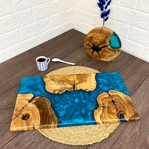 May include: A rectangular wooden serving board with a vibrant blue resin center. The board features natural wood grain and a hole for hanging. A small cup of coffee and a spoon are also present.