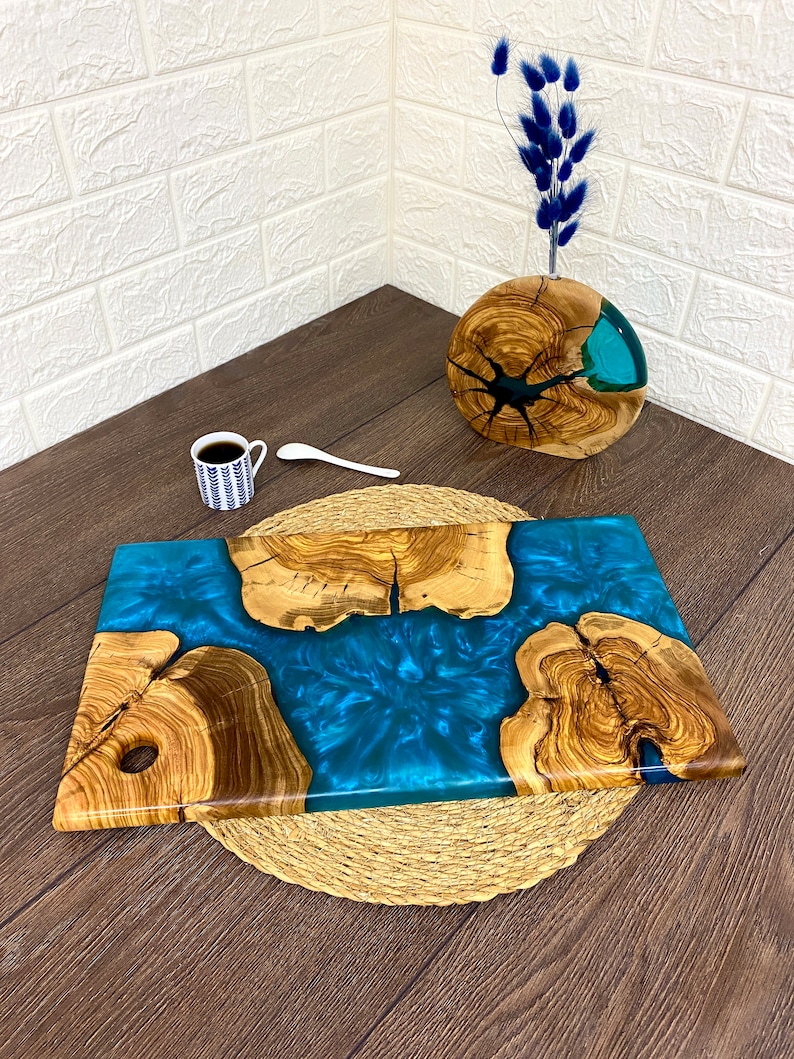 May include: A rectangular wooden serving board with a vibrant blue resin center. The board features natural wood grain and a hole for hanging. A small cup of coffee and a spoon are also present.