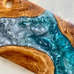 May include: A handcrafted wooden serving board with a river of teal and silver epoxy resin. The board features a natural wood grain border and a hole for hanging. A unique piece for serving food.