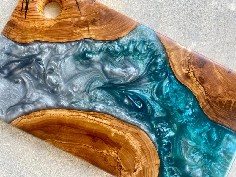 May include: A handcrafted wooden serving board with a river of teal and silver epoxy resin. The board features a natural wood grain border and a hole for hanging. A unique piece for serving food.