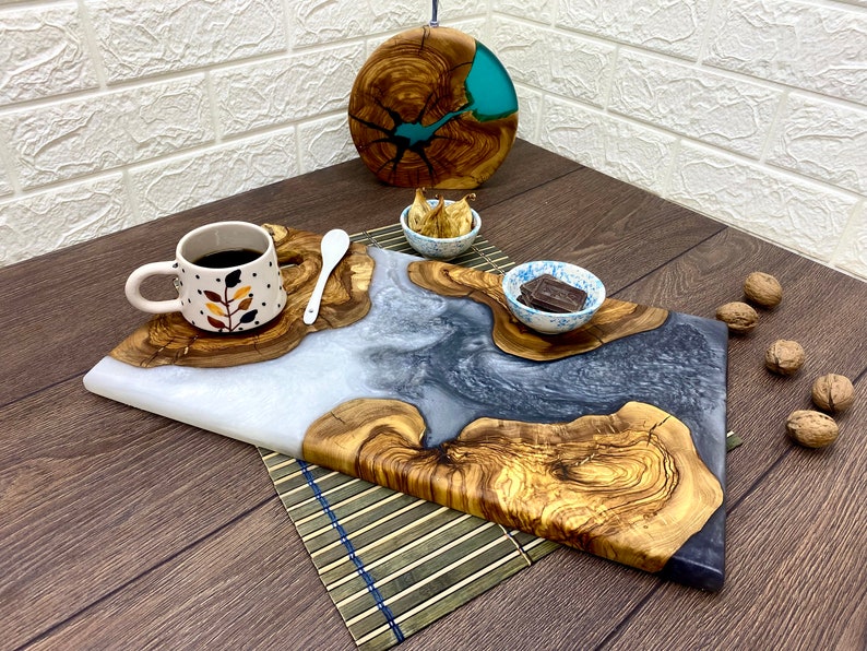 May include: A wooden serving board with a white and gray resin design, accompanied by a small cup of coffee, a spoon, and small bowls. A round wooden piece with blue resin is in the background. Walnuts are scattered around.