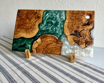 Puede incluir: Una tabla de cortar rectangular de madera con un diseño único. La tabla presenta secciones de vetas de madera natural, resina epoxi verde y blanca, y un agujero para colgar. Se muestra sobre un soporte de madera.