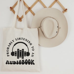 May include: A white canvas tote bag with black text that reads "Probably listening to an audiobook" with a graphic of headphones and books. The bag is hanging on a wooden rack.