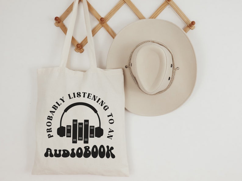 May include: A white canvas tote bag with black text that reads "Probably listening to an audiobook" with a graphic of headphones and books. The bag is hanging on a wooden rack.