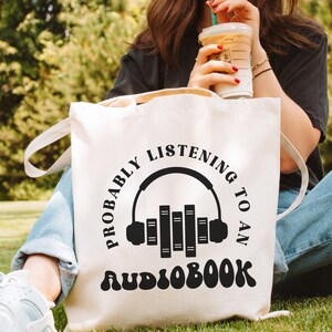 May include: White canvas tote bag with black text that reads "Probably listening to an audiobook" with a graphic of headphones and books.