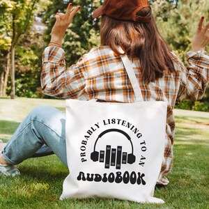 May include: A white tote bag with a black graphic that says "Probably listening to an audiobook" with a stylized image of headphones and books.