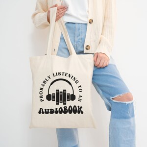 May include: A white canvas tote bag with black text that reads "Probably listening to an audiobook" and a graphic of headphones with books stacked inside.