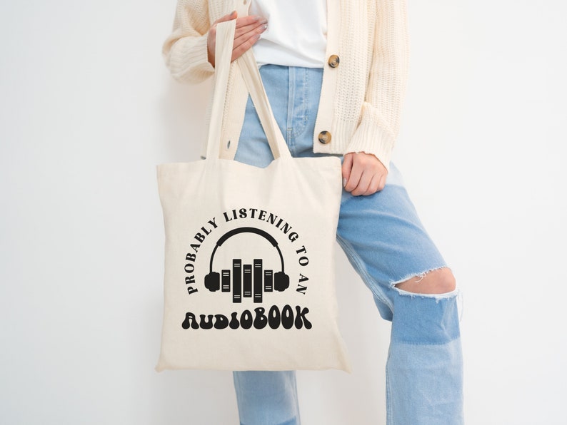 May include: A white canvas tote bag with black text that reads "Probably listening to an audiobook" and a graphic of headphones with books stacked inside.