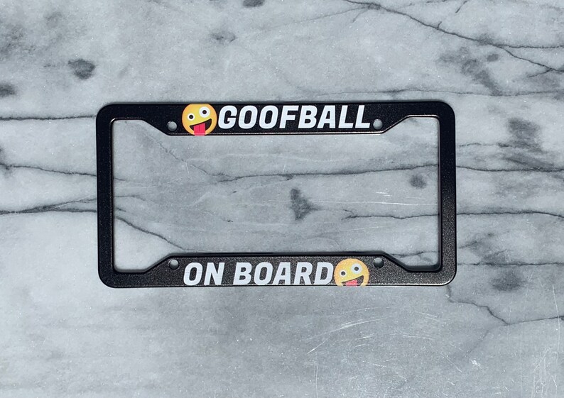 May include: Black license plate frame with white text that says "GOOFBALL" and a yellow smiley face with a red tongue. The bottom of the frame says "ON BOARD" with another yellow smiley face with a red tongue.