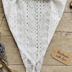 May include: White, triangular-shaped eyelet fabric with a scalloped edge. The fabric has a diamond pattern with small holes and floral lace details. A tag that says "Handmade with Love" is visible.
