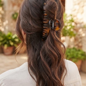 May include: A large, dark brown tortoiseshell hair claw clip with a glossy finish and a metal spring mechanism, securing a section of long, wavy, dark brown hair in a half-up style. The clip features a classic, elongated, multi-pronged design.