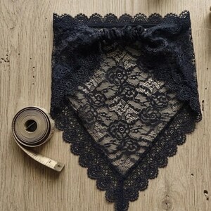 May include: A black triangular lace bandana featuring a floral pattern with scalloped edges and a gathered top band. A coiled beige measuring tape is positioned next to the lace accessory to provide a clear visual reference for its scale.