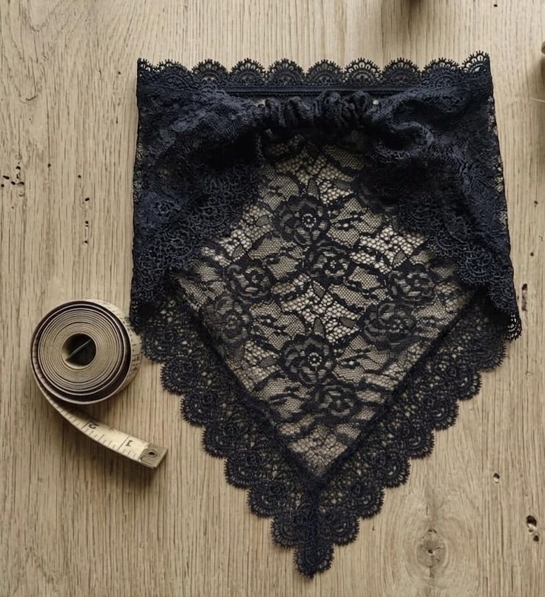 May include: A black triangular lace bandana featuring a floral pattern with scalloped edges and a gathered top band. A coiled beige measuring tape is positioned next to the lace accessory to provide a clear visual reference for its scale.