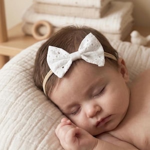 May include: A sleeping baby wearing a white eyelet bow headband. The bow is centered on a beige elastic band. The baby is resting on a cream-colored, textured pillow. Soft focus background with folded blankets and a wooden toy.