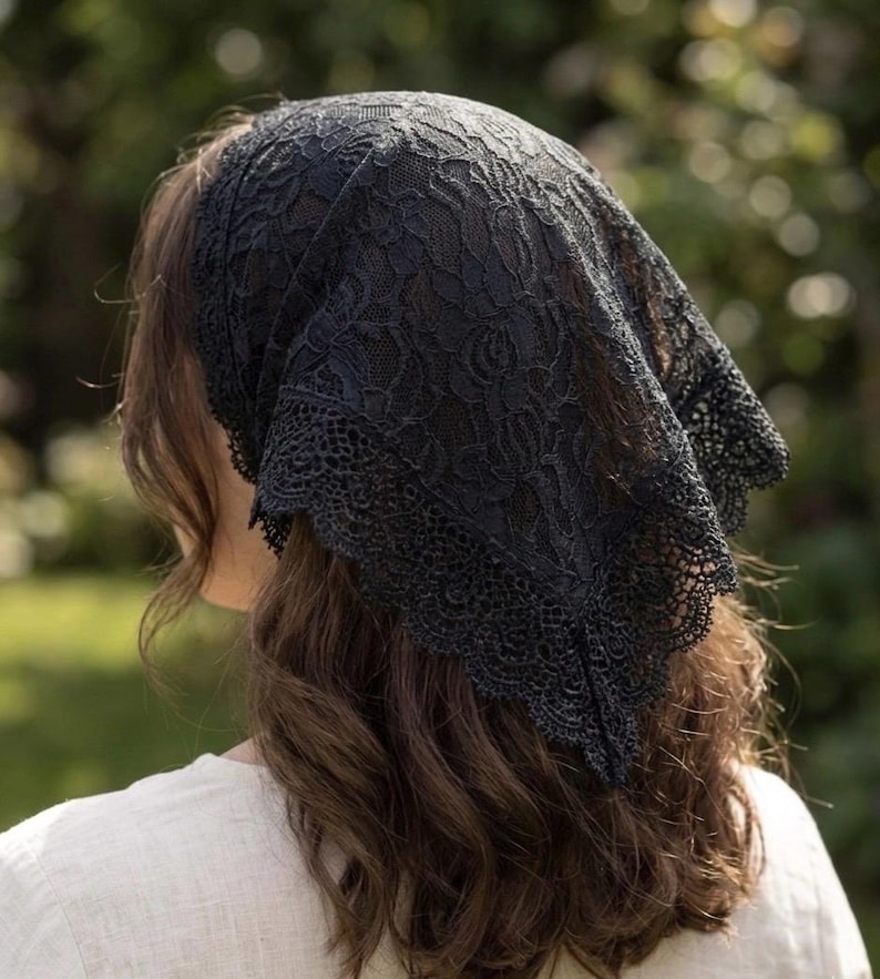 May include: A black lace head covering featuring a floral pattern and a scalloped edge. The triangular fabric piece is draped over the top of the head, with the intricate lace design covering the hair and extending down to the nape of the neck.