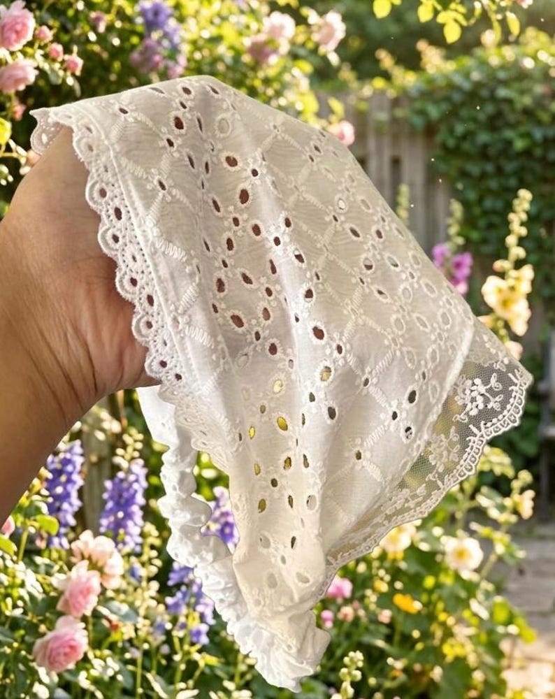 May include: White eyelet fabric with a scalloped lace edge. The fabric has a geometric pattern of small, evenly spaced holes. The background is a garden with pink and purple flowers.