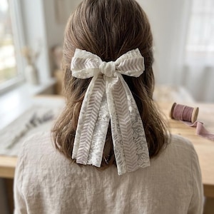 Könnte beinhalten: Eine Haarschleife aus weißer floraler Spitze und einem hellbeigen Band mit Chevron-Muster. Die Schleife ist in der Mitte geknotet und hat zwei lange, herabhängende Bänder, die die geschichtete Textur des Stoffes zur Geltung bringen.