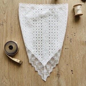 May include: White eyelet handkerchief with a diamond pattern and a delicate lace trim. A vintage measuring tape and a wooden spool of thread are visible on a wooden surface. The handkerchief is folded in a triangle shape.