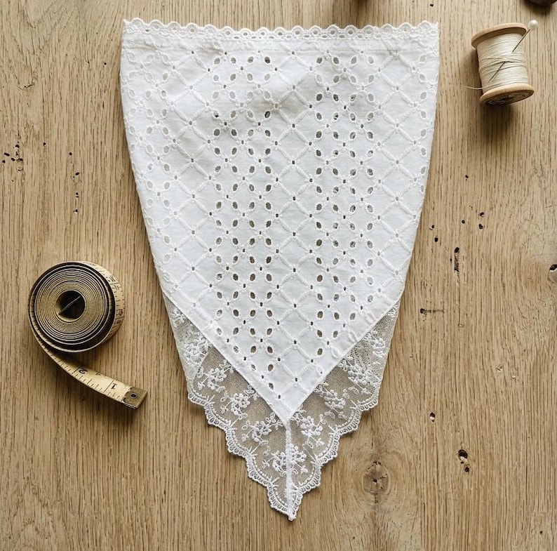 May include: White eyelet handkerchief with a diamond pattern and a delicate lace trim. A vintage measuring tape and a wooden spool of thread are visible on a wooden surface. The handkerchief is folded in a triangle shape.