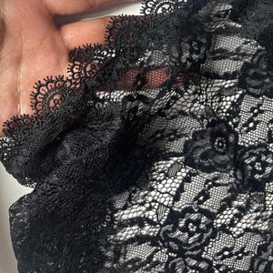 May include: A black lace fabric featuring a detailed floral pattern and a scalloped edge with intricate needlework. The material is semi-sheer, showcasing a delicate mesh construction with woven rose motifs throughout the entire textile surface.