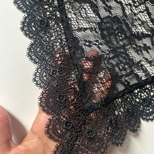 May include: A black lace fabric featuring a detailed floral pattern and a scalloped edge with intricate needlework. The material is semi-sheer with a fine mesh base, showcasing a delicate and ornate design throughout the textile surface.