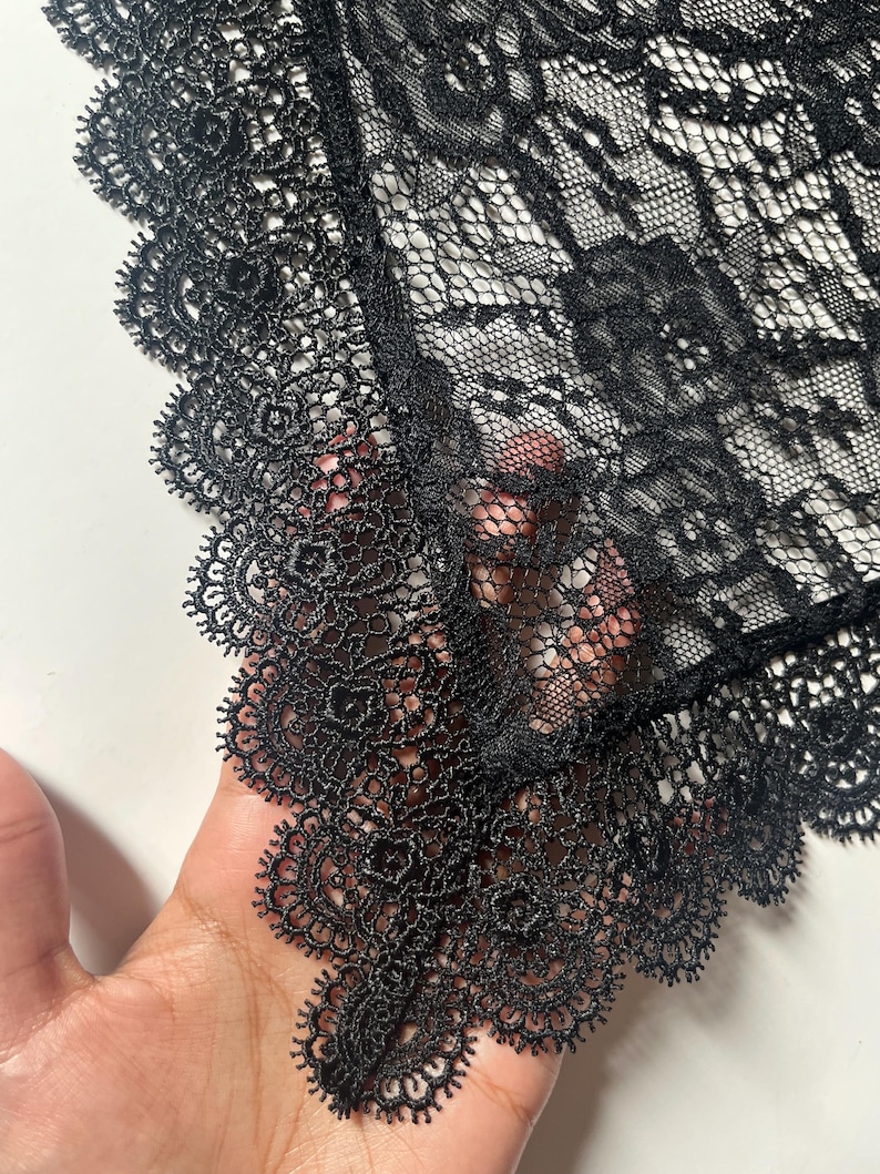 May include: A black lace fabric featuring a detailed floral pattern and a scalloped edge with intricate needlework. The material is semi-sheer with a fine mesh base, showcasing a delicate and ornate design throughout the textile surface.