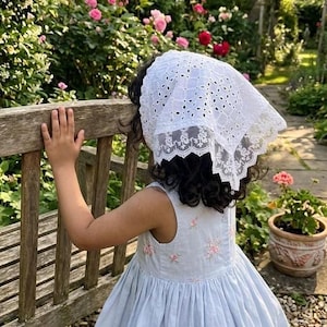 May include: A young person wearing a light blue dress with pink floral embroidery and a white eyelet lace bonnet. The scene is set in a garden with a wooden bench and blooming pink roses.