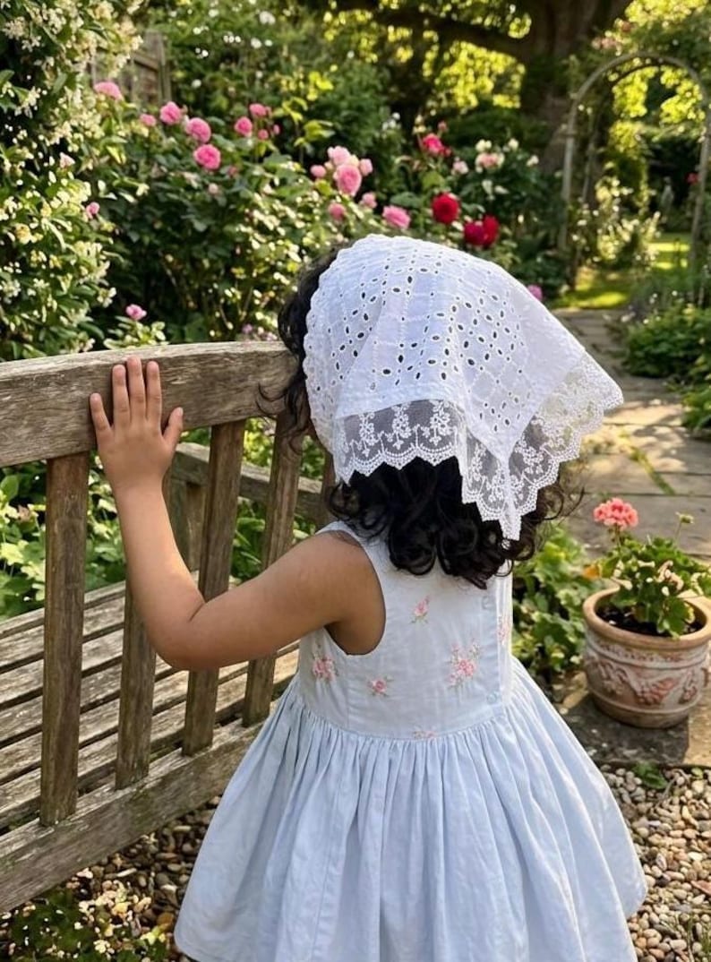 May include: A young person wearing a light blue dress with pink floral embroidery and a white eyelet lace bonnet. The scene is set in a garden with a wooden bench and blooming pink roses.