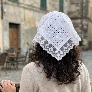 May include: A white eyelet lace head covering with a scalloped lace trim. The head covering is square-shaped and has a geometric pattern of small cut-out holes. The person is wearing a beige sweater.