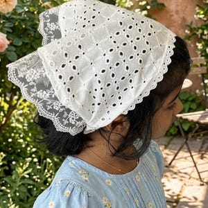 May include: A white eyelet bonnet with a scalloped lace trim. The bonnet is worn over dark hair, paired with a light blue dress with a daisy pattern. The background features a garden with pink roses and greenery.